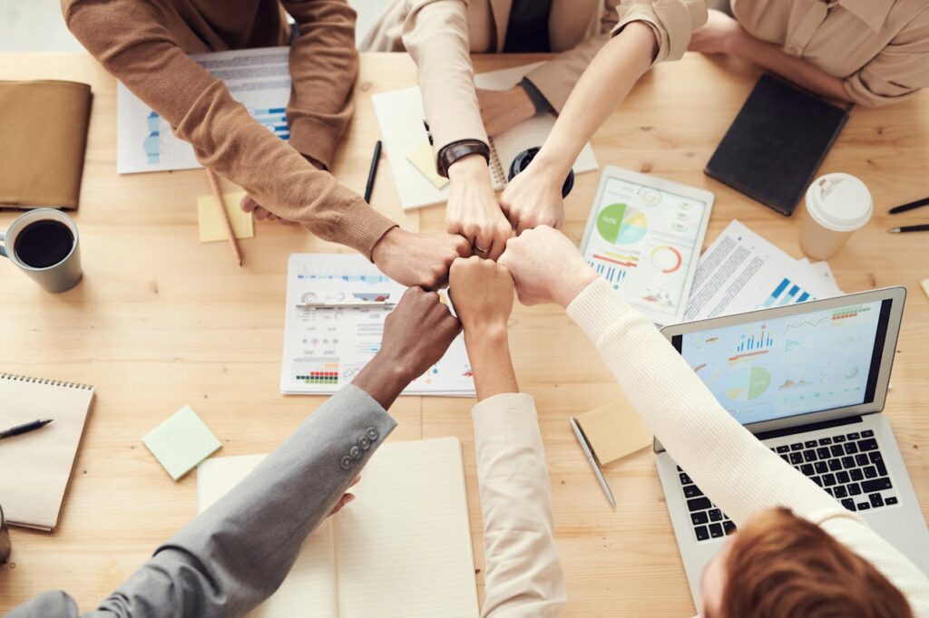 pexels-photo-3184438 Top view of a diverse team fist bumping over a meeting table with paperwork and laptops, symbolizing teamwork.